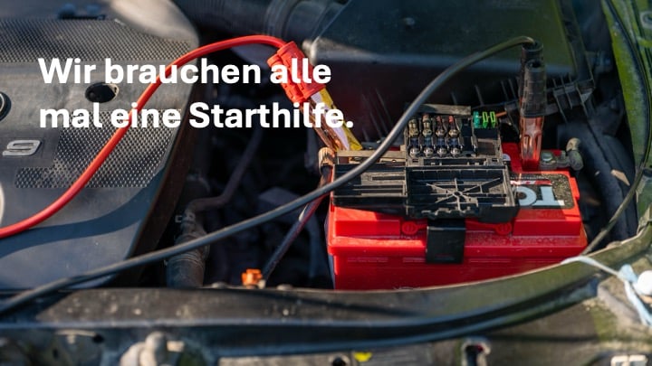 Starthilfekabel an einer Autobatterie mit dem Schriftzug „Wir brauchen alle mal eine Starthilfe.“